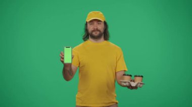 Kurye hizmeti konsepti. Elinde kahve tutacağı ve akıllı telefon tutan sarı şapkalı bir teslimatçının portresi. Paket servis ürünleri. Yeşil arka planda izole edilmiş. Reklam alanı, çalışma alanı modeli.