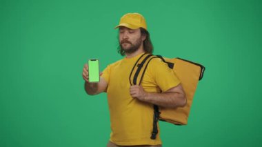 Sarı şapkalı ve tişörtlü bir teslimatçının portresi akıllı telefon ve portatif buzdolabı tutuyor. Yeşil arka planda izole edilmiş. Reklam alanı, çalışma alanı modeli.