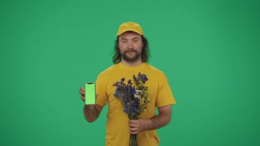 Kurye hizmeti konsepti. Sarı şapkalı ve tişörtlü bir teslimatçının portresi elinde bir buket çiçek ve akıllı telefon tutuyordu. Yeşil arka planda izole edilmiş. Reklam alanı, çalışma alanı modeli.