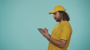 Kurye hizmeti konsepti. Sarı şapkalı bir teslimatçının portresi elinde tablet, içine bir şeyler yazıyor, kameradan başka yere bakıyor. Düzen kurmak. Mavi arkaplanda izole.