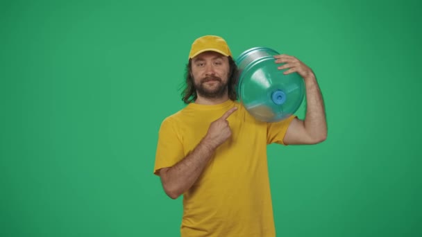 快递服务的概念 一个头戴黄帽 身穿T恤的送货人的画像 肩上扛着大水瓶 指着空旷的地方 广告空间 在绿上被隔离 — 图库视频影像