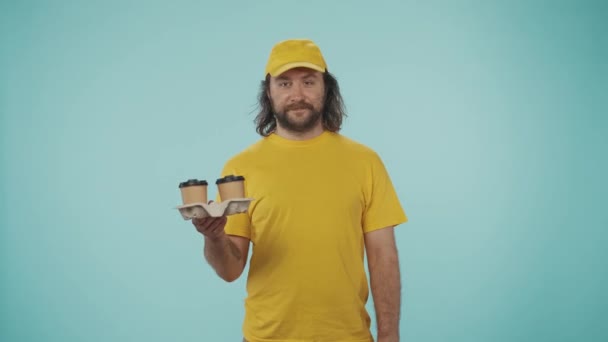 宅配サービスのコンセプト コーヒーカップホルダーと紙袋を持っている黄色い帽子の友好的な配達男性の肖像画 カフェテイクアウト製品 青い背景に孤立した — ストック動画