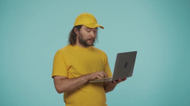快递服务的概念 一个头戴黄色帽子的送货员拿着笔记本电脑 在里面输入一些东西 并对着摄像机说话的画像 订单信息 与蓝色背景隔离 — 图库视频影像