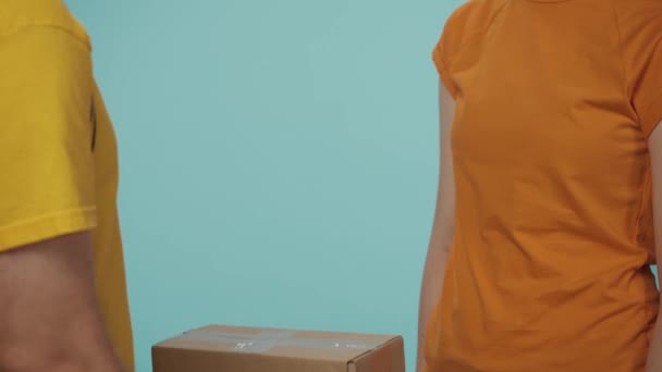 クローズアップショット 宅配サービスのコンセプト 黄色いTシャツの配達男性の肖像画は 彼女の注文したパッケージを女性の顧客に与え 少女はタブレットで注文にサインします 青い背景に孤立した — ストック動画