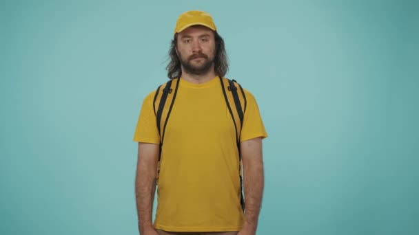 宅配サービスのコンセプト カメラにポーズするポータブルバックパック冷蔵庫で黄色い帽子の配達男性の肖像画 青い背景に孤立した — ストック動画