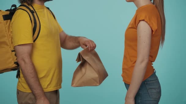 快递服务的概念 一个头戴黄色帽子头戴便携背包的送货男子的肖像给了一个女顾客纸袋和她的食品订单 与蓝色背景隔离 — 图库视频影像