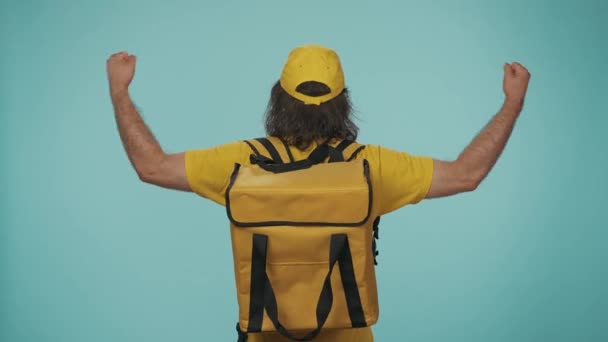 快递服务的概念 一个带着手提式背包冰箱头戴黄色帽子 背对着摄像机举手表决的送货员的画像 与蓝色背景隔离 — 图库视频影像
