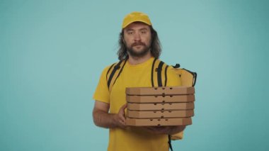 Kurye hizmeti konsepti. Sırt çantalı, portatif buzdolabı taşıyan sarı şapkalı bir teslimatçının portresi bir yığın pizza kutusunu tutuyor ve onaylıyor. Mavi arkaplanda izole.