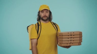 Kurye hizmeti konsepti. Sırt çantalı, portatif buzdolabı taşıyan sarı şapkalı bir teslimatçının portresi bir yığın pizza kutusu tutuyor ve tamam işareti gösteriyor. Mavi arkaplanda izole.