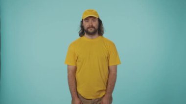 Kurye hizmeti konsepti. Sarı şapkalı ve tişörtlü bir teslimatçının portresi. Elinde bir kese kağıdıyla kameraya veriyor. Mavi arkaplanda izole.