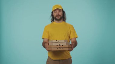 Kurye hizmeti konsepti. Sarı şapkalı ve tişörtlü bir teslimatçının portresi bir yığın pizza kutusunu tutuyor ve aç yüz ifadesi ve jestleriyle ne kadar lezzetli olduğunu gösteriyor. Mavi üzerine izole edilmiş.