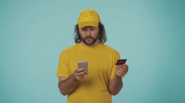 Sarı şapkalı ve tişörtlü bir teslimatçının portresi banka kartı tutuyor ve akıllı telefonuna numara yazıyor. Çevrimiçi ödeme kavramı. Mavi arkaplanda izole.