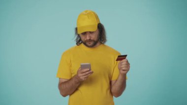 Elinde banka kartı ve akıllı telefon tutan sarı şapkalı ve tişörtlü bir teslimatçının portresi. İnternetten bir şey alan adam. Ödeme konsepti. Mavi arkaplanda izole.