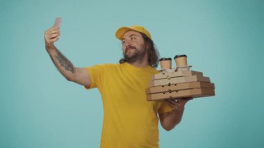 Kurye hizmeti konsepti. Elinde pizza kutuları ve kahve tutacağı olan sarı şapkalı bir teslimatçının portresi. Selfie çekiyor ve video kaydediyor. Sosyal medya paylaşımı. Mavi arkaplanda izole.