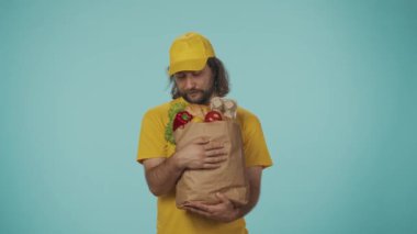 Gıda piyasası kurye hizmeti konsepti. Sarı şapkalı bir teslimatçının portresi. Elinde alışveriş ürünleriyle dolu büyük bir kesekağıdı var. Kameraya bakıyor. Mavi arkaplanda izole.