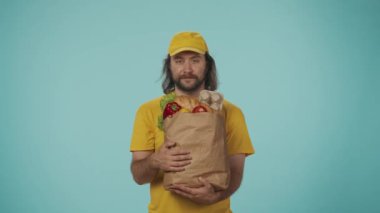 Gıda piyasası kurye hizmeti konsepti. Sarı şapkalı bir teslimatçının portresi. Elinde yiyecek ürünleriyle dolu büyük bir kese kağıdı var. Onaylıyor. Mavi arkaplanda izole.