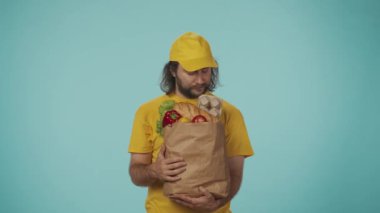 Gıda piyasası kurye hizmeti konsepti. Sarı şapkalı bir teslimatçının portresi market ürünleriyle dolu büyük çantaya bakıyor, baş parmağını kaldırıyor, gülümsüyor. Mavi arkaplanda izole.