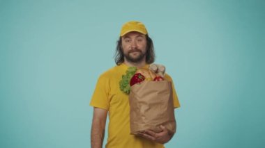 Gıda piyasası kurye hizmeti konsepti. Sarı şapkalı bir teslimatçının portresi bir elinde market ürünleriyle dolu bir çanta ve baş parmak işareti gösteriyor. Mavi arkaplanda izole.