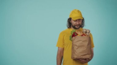 Gıda piyasası kurye hizmeti konsepti. Sarı şapkalı bir teslimatçının portresi market ürünleriyle dolu bir çanta tutuyor ve boş alanı işaret ediyor. Reklam için yer var. Mavi arkaplanda izole.