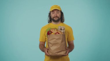 Gıda piyasası kurye hizmeti konsepti. Sarı şapkalı bir teslimatçının portresi elinde alışveriş ürünleriyle dolu büyük bir kesekağıdı ile kameraya poz veriyor. Mavi arkaplanda izole.