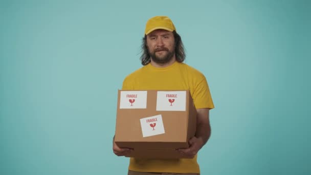 宅配サービスのコンセプト パッケージの上に置かれた壊れやすいステッカーが付いている非常に重い箱を握っている黄色い帽子の配達人の肖像画 青い背景に孤立した — ストック動画
