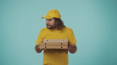 Kurye hizmeti konsepti. Sarı şapkalı ve tişörtlü bir teslimatçının portresi bir yığın pizza kutusunu tutuyor ve aç yüz ifadesi ve jestleriyle ne kadar lezzetli olduğunu gösteriyor. Mavi üzerine izole edilmiş.