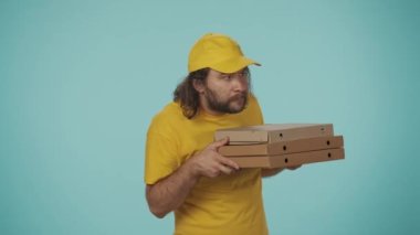 Kurye hizmeti konsepti. Sarı şapkalı ve tişörtlü bir teslimatçının portresi pizza kutularını tutuyor, etrafa bakıyor, açıyor ve bir ısırık alıyor, tadını çıkarıyor. Mavi arkaplanda izole.