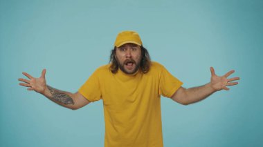Kurye hizmeti konsepti. Sarı şapkalı ve tişörtlü bir teslimatçının portresi kameraya bakıyor, akıl almaz bir jest ifadesi yapıyor. Mavi arkaplanda izole.