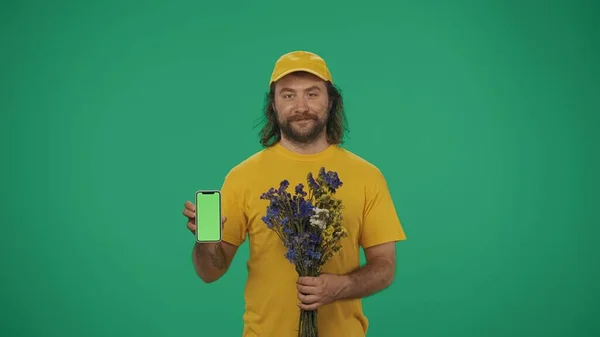 Portret Kuriera Żółtej Czapce Bukietem Kwiatów Smartfonem Dłoni Odizolowany Zielonym — Zdjęcie stockowe