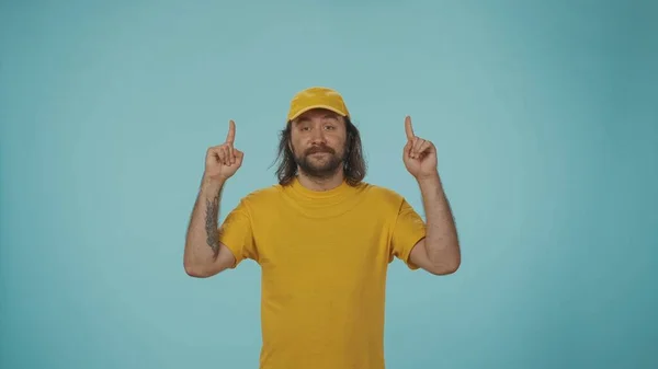 快递服务的概念 一个头戴黄色帽子 身穿黄色T恤的送货人的画像 手指指向上方 在镜头前摆姿势 做广告的地方与蓝色背景隔离 — 图库照片