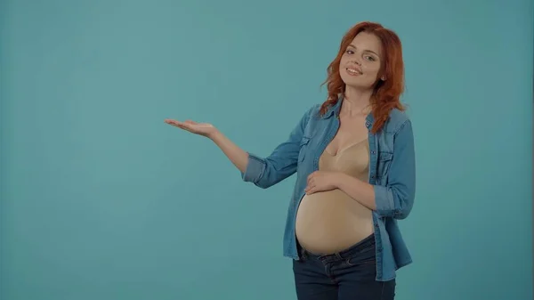 一个红头发的女人怀了孩子 抱着怀孕的肚子 用手掌指向一个空的地方做广告 一个蓝色背景的孕妇在工作室里 — 图库照片