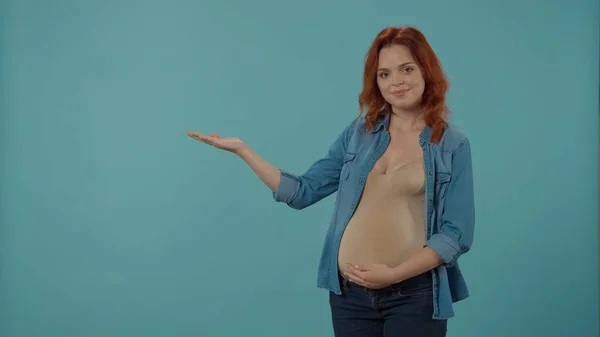 一个红头发的女人怀了孩子 抱着怀孕的肚子 用手掌指向一个空的地方做广告 一个蓝色背景的孕妇在工作室里 — 图库照片