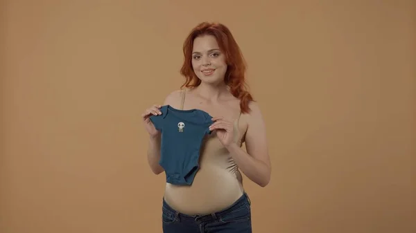 青い赤ちゃんのボディスーツを持っている妊婦は 妊娠中に男の子の誕生を準備しています オレンジ色の背景にあるスタジオの若い母親 女は新生児を待っている コンセプト — ストック写真