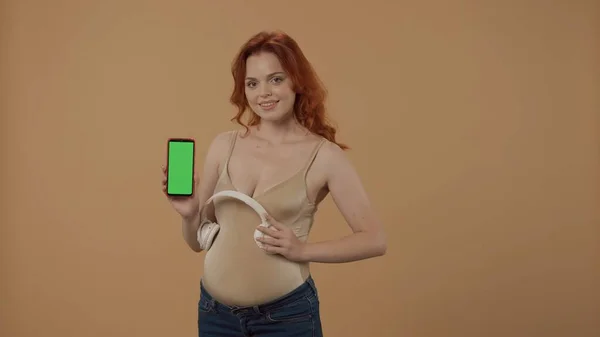 妊娠中の女性は 白いワイヤレスヘッドフォンを胃に置き 彼女の手に緑色のスクリーンが付いているスマートフォンを保持します オレンジ色の背景にあるスタジオの女性 Aの開発の概念 — ストック写真