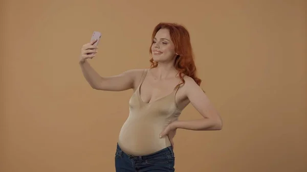 ハッピータイム 赤毛の妊婦がセルフィーを取って スマートフォンを使ってビデオ通話をする オレンジ色の背景にあるスタジオで妊娠した女性 母性という概念 — ストック写真