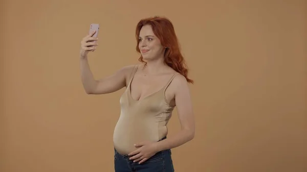 ハッピータイム 赤毛の妊婦がセルフィーを取って スマートフォンを使ってビデオ通話をする オレンジ色の背景にあるスタジオで妊娠した女性 母性という概念 — ストック写真