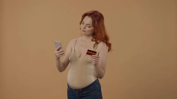 オンライン決済 妊娠中の女性は スマートフォンを使用してクレジットカードでオンラインストアで購入します オレンジ色の背景にあるスタジオで妊娠した女性 母性という概念 — ストック写真