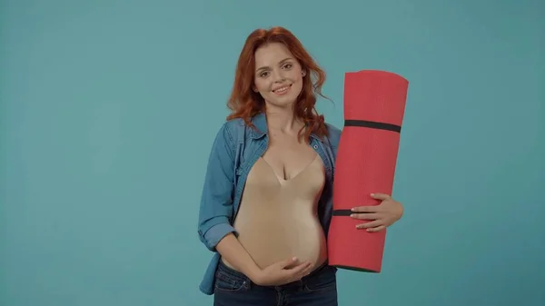 青い背景にスタジオでヨガマットを保持している若い妊婦の肖像画 女性は妊娠中に健康でアクティブなライフスタイルをリードします スポーツ 妊娠の概念 — ストック写真
