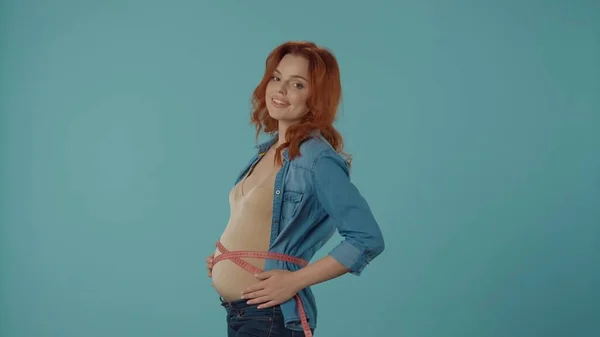 ポジティブな妊娠中の女性は 青い背景に立ってテープと笑顔で彼女の腹を測定します 健康な妊娠 産婦人科 — ストック写真