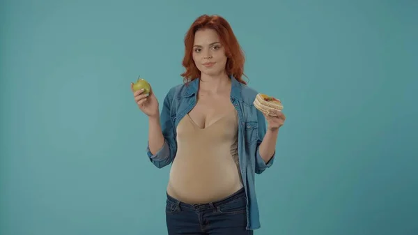 妊娠中の女性は健康で不健康な食べ物を選ぶことはできません 女性は甘いケーキと緑のリンゴの間で選択します 青い背景にあるスタジオの女性 — ストック写真