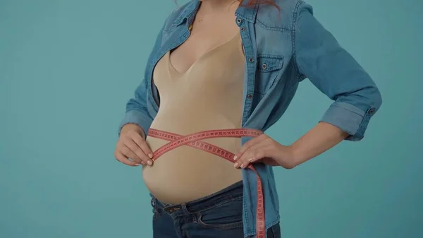 妊娠中の女性は 青い背景に立ってテープで腹を測定します 妊娠中の女性の血まみれの腹が閉じ込められています 妊娠中のサイズと体のパラメータをチェックする — ストック写真