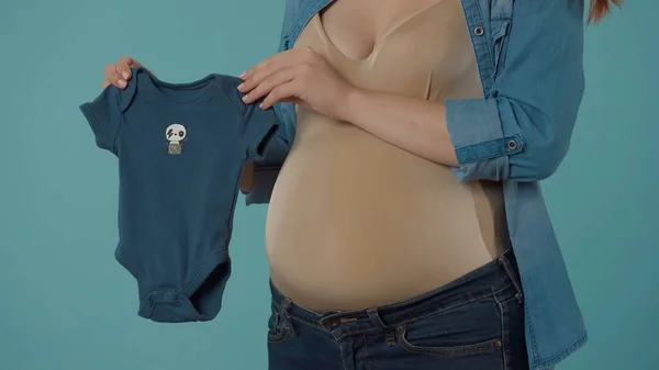 一名穿着蓝色婴儿紧身衣的孕妇正在为怀孕期间生男孩做准备 在一个蓝色背景的工作室里 一个怀孕妇女的腹部肿胀 那个女人 — 图库照片