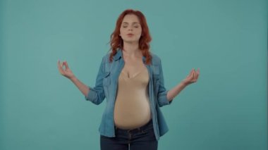 Kızıl saçlı hamile kadın stüdyoda meditasyon yapıyor. Mavi arka planda. Kadın ellerini bir Mudra hareketiyle gözlerini kapatmış ve derin bir nefes alıp veriyor.