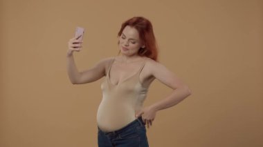Kızıl saçlı hamile kadın bir video konuşmasında akıllı telefon kullanıyor. Bir kadın selamlaşırken elini sallıyor, koca göbeğini gösteriyor ve veda ediyor. Stüdyoda turuncu arka planda hamile bir kadın var.