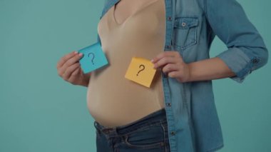 Hamile bir kadın, göbeğinde soru işaretleri olan kare kağıtlar tutuyor. Stüdyoda, mavi arka planda, hamile bir kadının göbeği şişmişti. Doğumu bekleniyor.