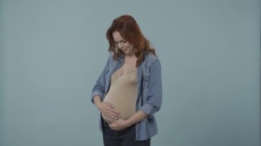 Genç hamile bir kadın şişmiş karnını sevgi ve şefkatle okşar. Stüdyoda mavi arka planda hamile bir kadının portresi. Hamilelik, annelik ve aşk kavramı.