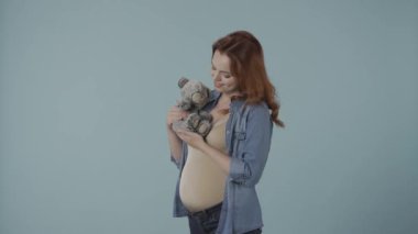 Kızıl saçlı hamile kadın elinde oyuncak ayı tutuyor. Stüdyoda elinde oyuncak olan mavi arka planda bir kadın. Duygusal sevgi dolu hamilelik zamanı. Bebek beklentisi. Sevgi ve ilgi