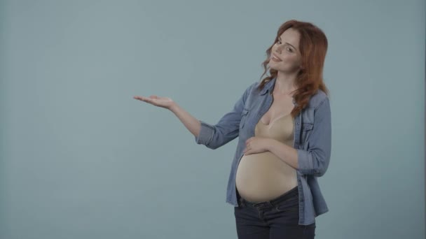 赤ちゃんを待っている女性は 妊娠中の腹を抱えて 広告のための空のスペースに彼女の手のひらを指します 妊娠中の女性は 青い背景にあるスタジオで彼女の頭を承認します コンセプト — ストック動画
