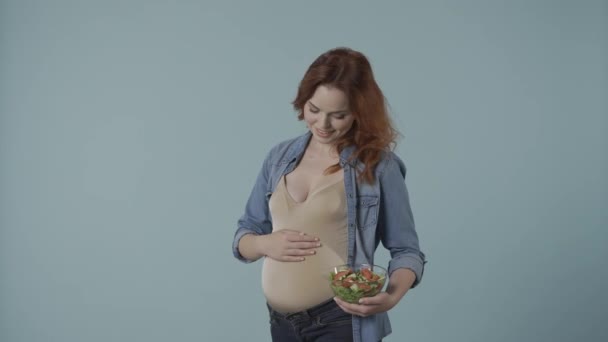 一个孕妇拿着一个透明的玻璃盘子 上面放着沙拉 指着它 然后竖起一个大拇指 一个蓝色背景的女人在工作室里孕妇的健康和适当营养 — 图库视频影像