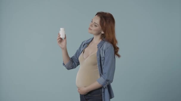 青い背景にスタジオでビタミン瓶を塗った妊婦のプロフィール写真 ビタミンを指差して親指を示す女性 妊娠期間中の医療と出生前のビタミン — ストック動画
